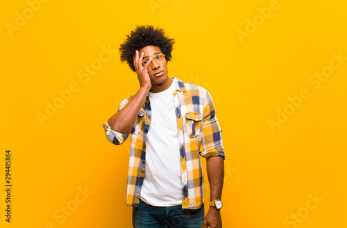 young black man feeling bored, frustrated and sleepy after a tiresome, dull and tedious task, holding face with hand against orange wall