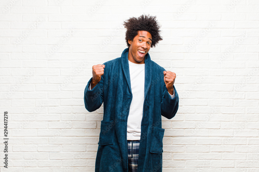 young black man wearing pajamas with gown feeling shocked, excited and happy, laughing and celebrating success, saying wow! against brick wall - obrazy, fototapety, plakaty 