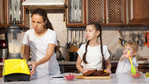 Mom is tasting and cooking cream for cake using blender with her two daughters in kitchen at home, girls are looking at process with interest, little girl is eating berries with culinary spatula. photo