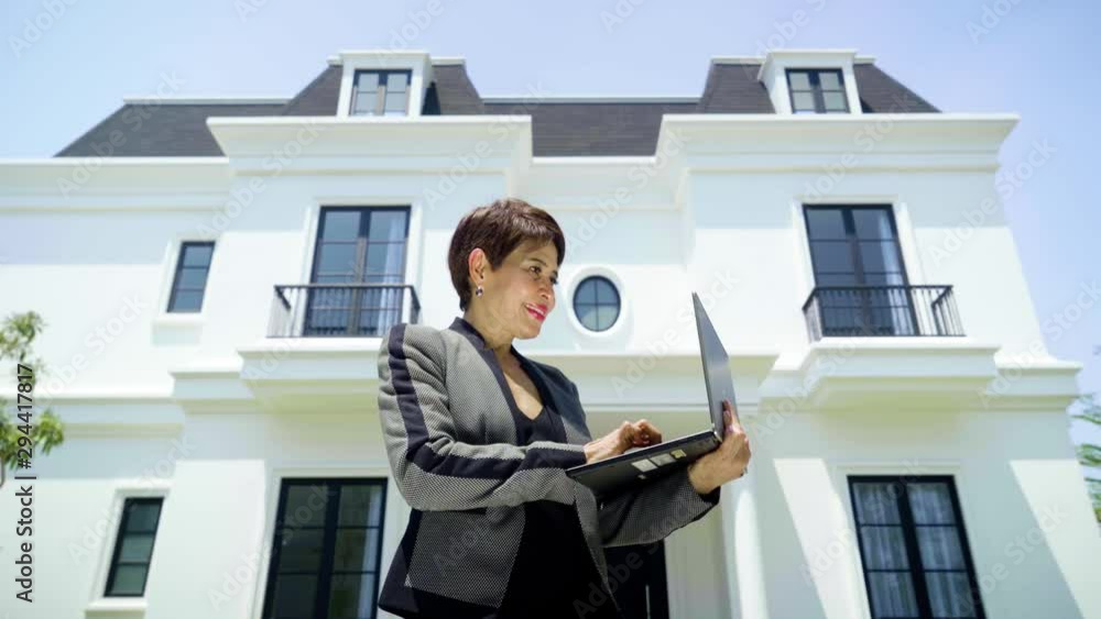 Female real estate working with a laptop computer while standing in front of modern house and smiling at the camera. Shot in 4k resolution Female real estate working with a laptop computer while standing in front of modern house and smiling at the camera. Shot in 4k resolution
