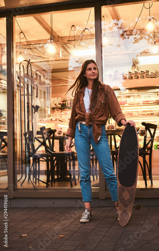Pretty young woman is standing near cafe while holding her longboard.