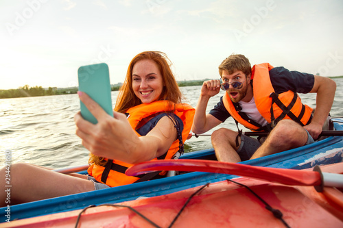 Happy young caucasian couple kayaking on river with sunset in the backgrounds. Having fun in leisure activity. Happy male and female model make selfie on the kayak. Sport, relations concept. Colorful. © master1305
