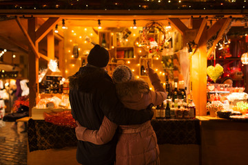 love, winter holidays and people concept - happy senior couple hugging at christmas market souvenir shop stall in evening