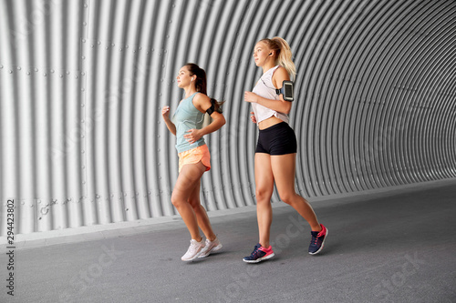 fitness, sport and healthy lifestyle concept - young women or female friends with earphones wearing armbands with smartphones and running outdoors