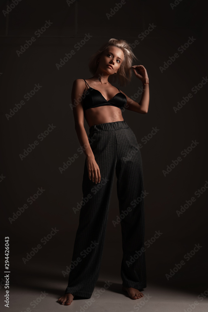 Stylish fashion trendy blonde girl with elegant hairstyle in classic trousers and black lingerie posing on black background. Beautiful hot sexy portrait of young woman model underwear in room Stylish fashion trendy blonde girl with elegant hairstyle in classic trousers and black lingerie posing on black background. Beautiful hot sexy portrait of young woman model underwear in room