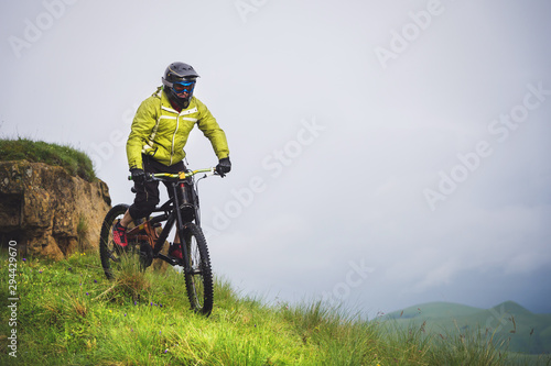 Fototapeta Naklejka Na Ścianę i Meble -  Front view of a man on a mountain bike standing on a rocky terrain and looking down against a gray sky. The concept of a mountain bike and mtb downhill