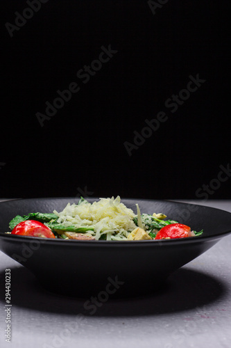 Fototapeta Naklejka Na Ścianę i Meble -  Concept for a tasty and healthy meal. Crispy bacon. Salad with chicken fillet with green cabbage. Big red cherry tomatoes. Black dish. Dark background. White table. Tender parmesan cheese.