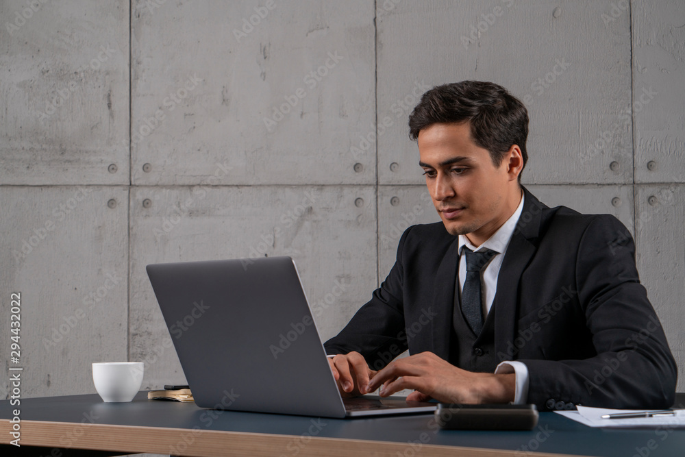 Handsome serious young businessman in dark suit using laptop computer in loft office. Concept of accounting and computer science Handsome serious young businessman in dark suit using laptop computer in loft office. Concept of accounting and computer science