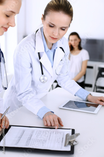 Group of doctors at work in hospital with patient in a queue at the background. Physician filling up medical documents or prescription while standing at reception desk. Data in medicine and health