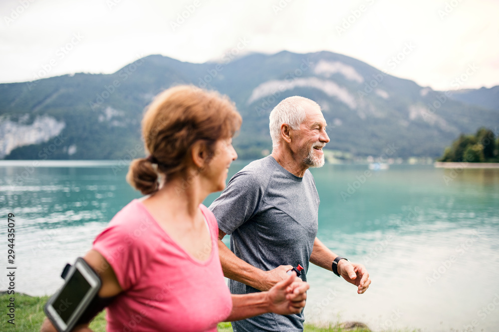 Fototapeta premium Senior pensioner couple with smartphone running by lake in nature.