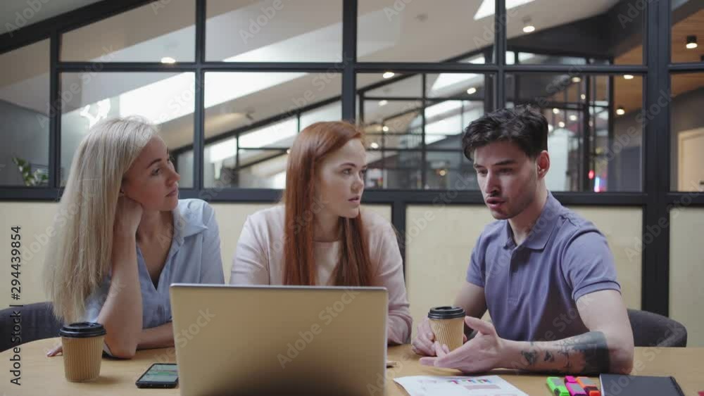 Camera tilting down to group of colleagues with coffee to go discussing project data on laptop while sitting at table in modern office Camera tilting down to group of colleagues with coffee to go discussing project data on laptop while sitting at table in modern office