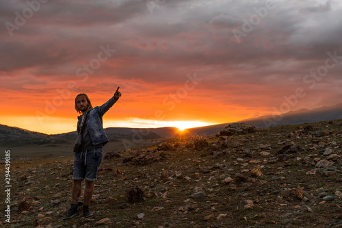A traveler stands in full growth at sunset in the mountains. Points a finger to the side. Backpacker in a hike in the summer mountains. A man in a denim jacket, shorts, long blond hair. Mountain Altai