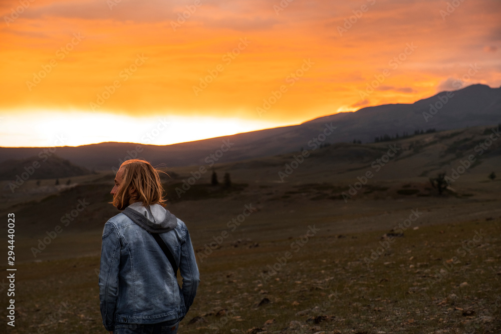 Sunset in the mountains. A man in a denim jacket, long blond hair. A tourist walks in the nature of Altai. The back of a man. The sun sets over the mountains, painting the sky in red and yellow.