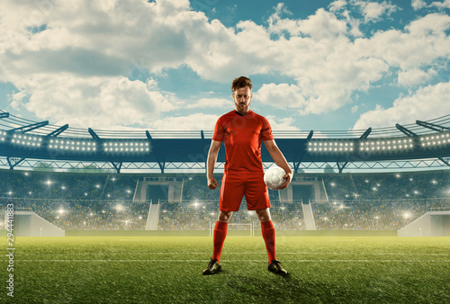 Professional soccer player in sports uniform with a ball on a field with tribunes, green grass and spectators. Blue cloudy sky © TandemBranding