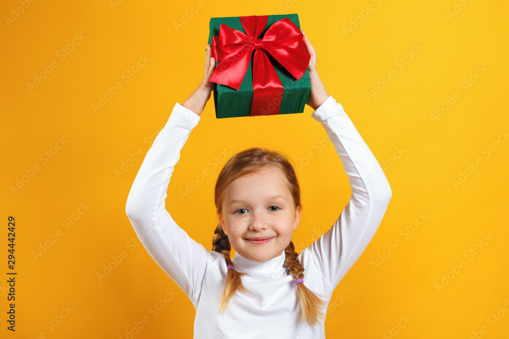 Cute happy little girl holding a box with a gift over her head. A child with pigtails in a white turtleneck on a yellow background Cute happy little girl holding a box with a gift over her head. A child with pigtails in a white turtleneck on a yellow background