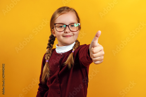 Little girl in glasses shows thumb up. A child in a burgundy sweater on a yellow background