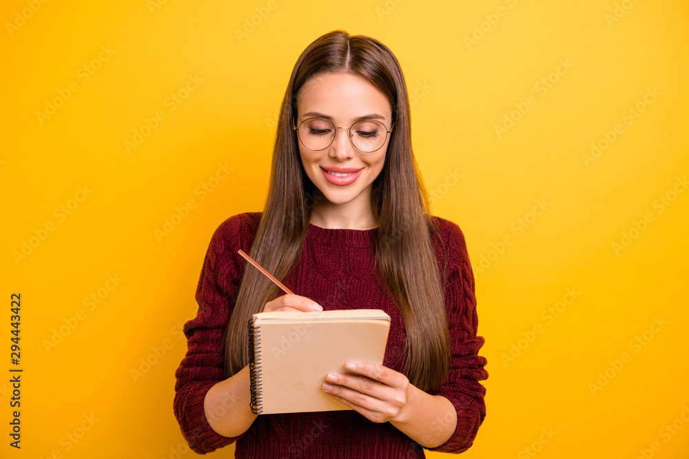 Portrait of charming girl holding pen and notebook writing check plan to do list wearing maroon jumper isolated over yellow background Portrait of charming girl holding pen and notebook writing check plan to do list wearing maroon jumper isolated over yellow background