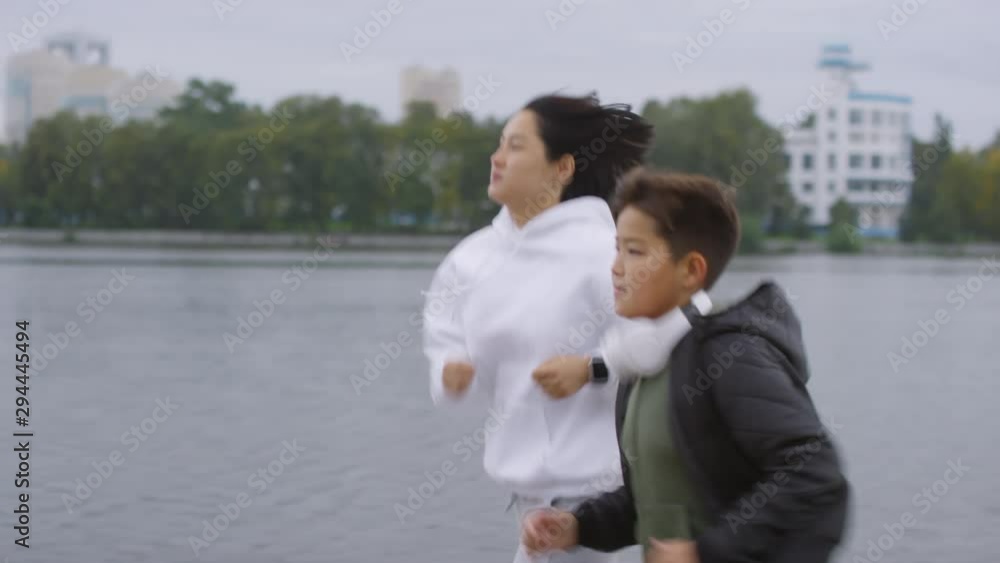 Side view of Asian mother and son in sportswear running along city riverside sidewalk in the morning Side view of Asian mother and son in sportswear running along city riverside sidewalk in the morning
