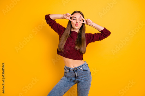 Portrait of lovely woman with eyes closed making v-signs wearing denim jeans burgundy jumper isolated over yellow background