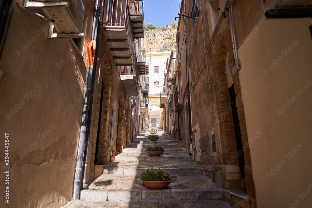 Typical example of historic mediterranean architecture of italian narrow small street in city Cefalu on the island Sicily, houses build from stones with balcony. Picture is taken in sunny spring day.