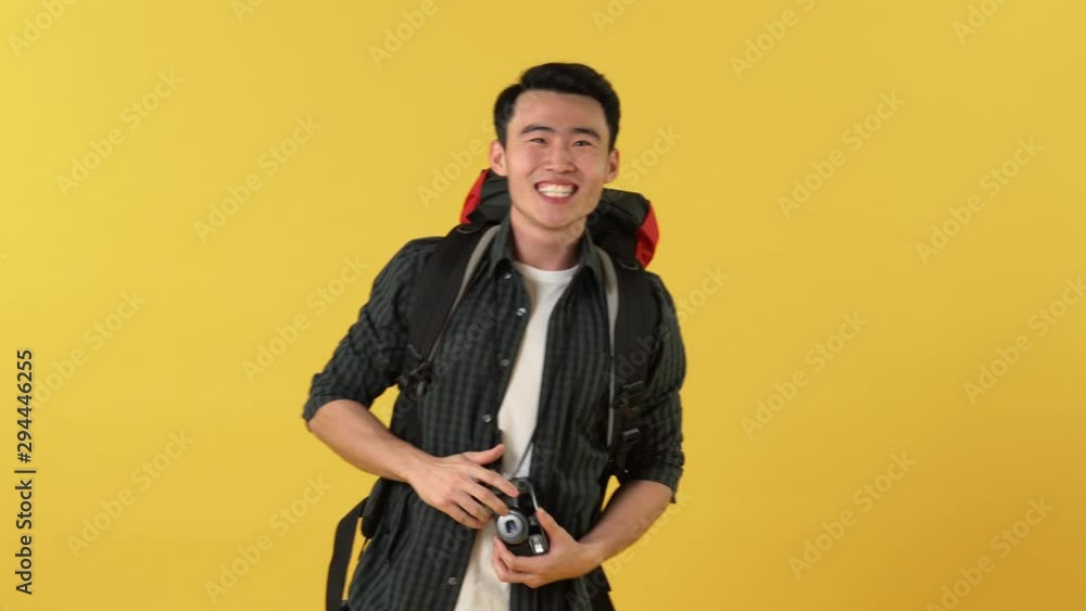 Young Asian tourist man with backpack feeling excited smiling and taking picture with camaera on yellow background Young Asian tourist man with backpack feeling excited smiling and taking picture with camaera on yellow background