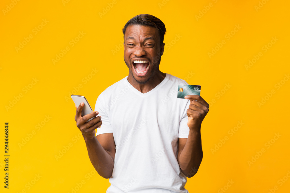 Afro Guy Holding Phone And Credit Card Shouting, Yellow Background Afro Guy Holding Phone And Credit Card Shouting, Yellow Background