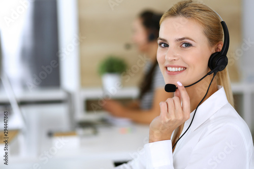 Blonde business woman using headset for communication and consulting people at customer service office. Call center. Group of operators at work at the background