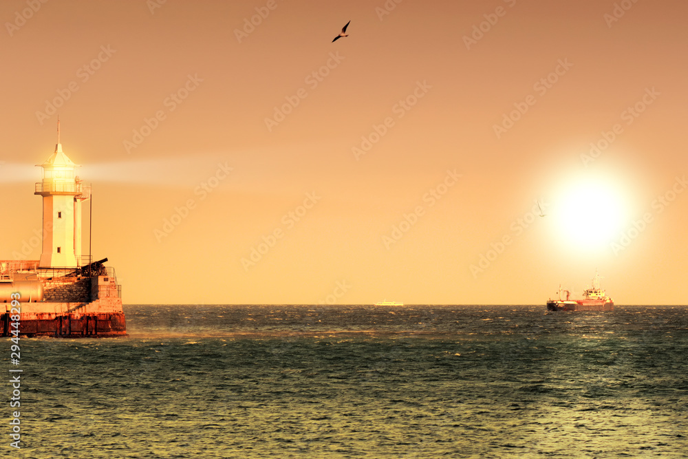 lighthouse at sunset with ship in sea water landscape against orange sky background wide view of ocean cargo vessel and navigation tower building in port scenic sunrise on shore