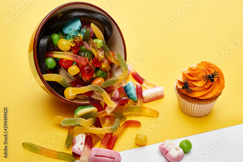 colorful gummy sweets scattered from bowl near cupcake on yellow background, Halloween treat