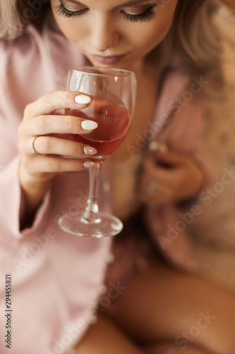 Modish sexy young confident woman, with long platinum blonde hair, in pink silk pajamas sitting in an armchair holding wine glass and drinking. Concept of brides morning before the wedding