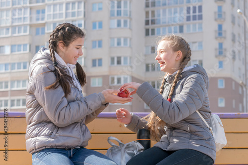 two white caucasian cute teenager funny girlfriends sitting together on bench sharing apple food