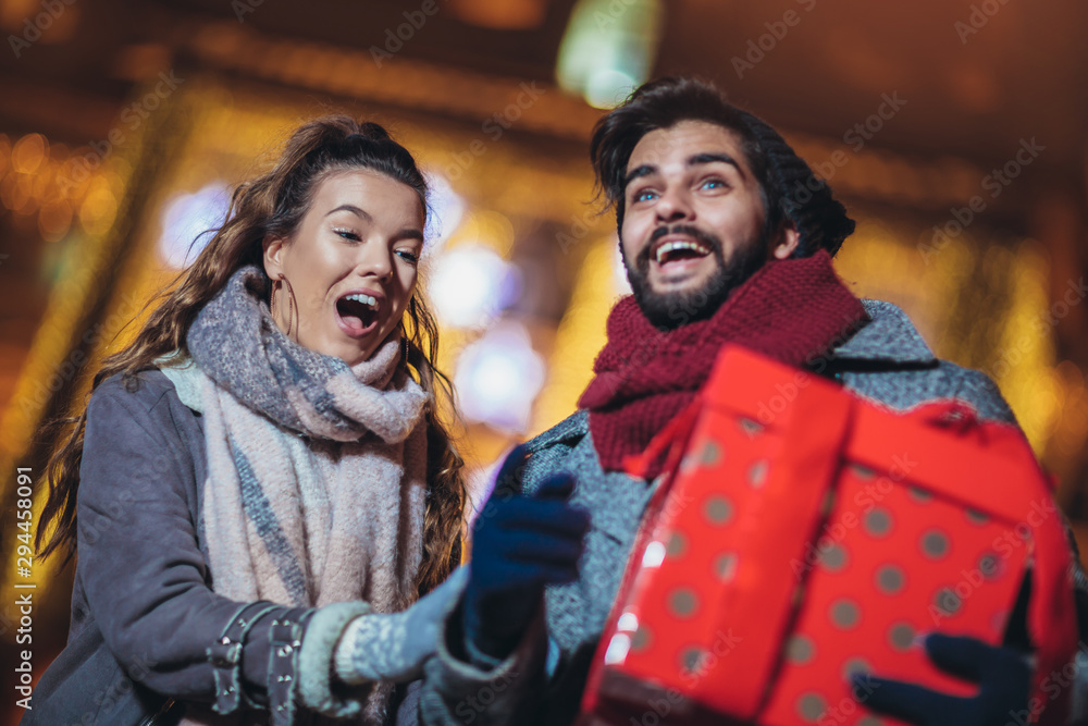Young couple in the city centre with holiday's brights in background. Man presenting gift to woman.