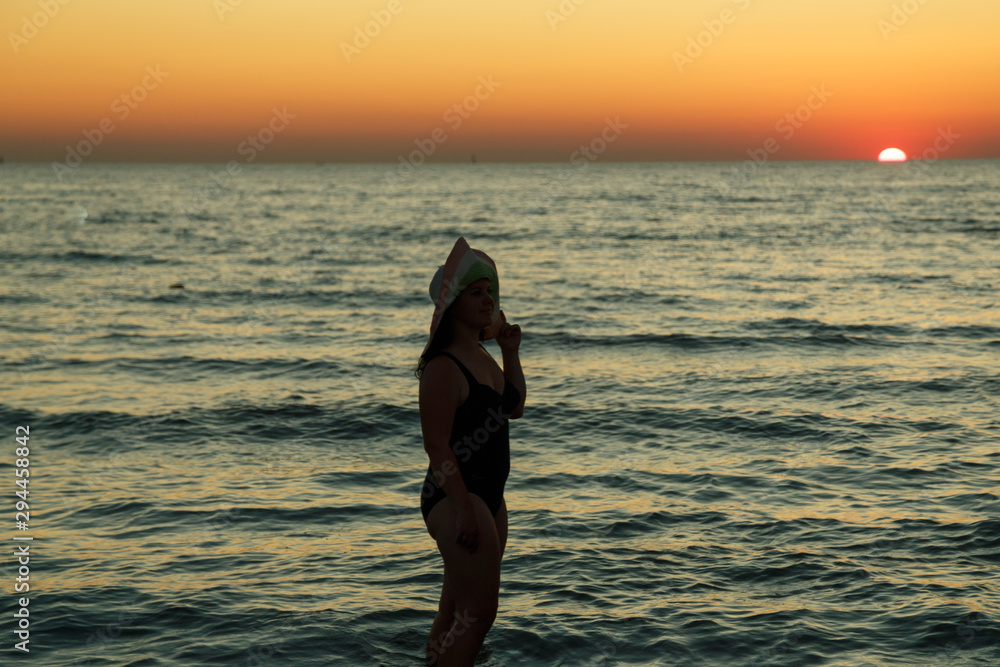 A woman in a hat walks along the seashore at dusk.
