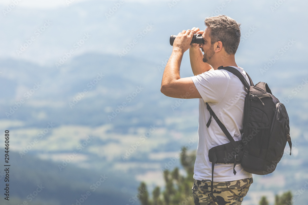 Traveler with backpack standing on the top of the mountain and using binoculars