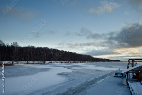 ice-covered river with a trace from the boat on it, a forest on the far shore, a snow-covered boat at the pier