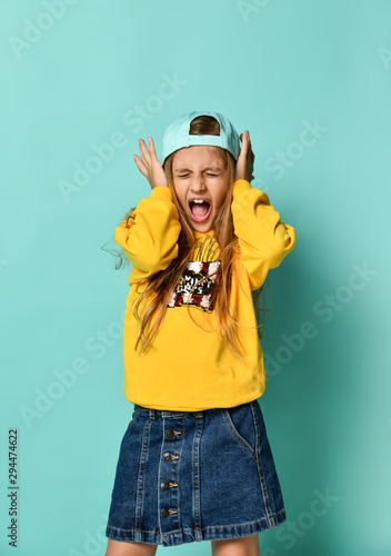 Pretty fashionable young teenager with a lovely smile posing in a baseball cap and yellow hoodie on a blue background
