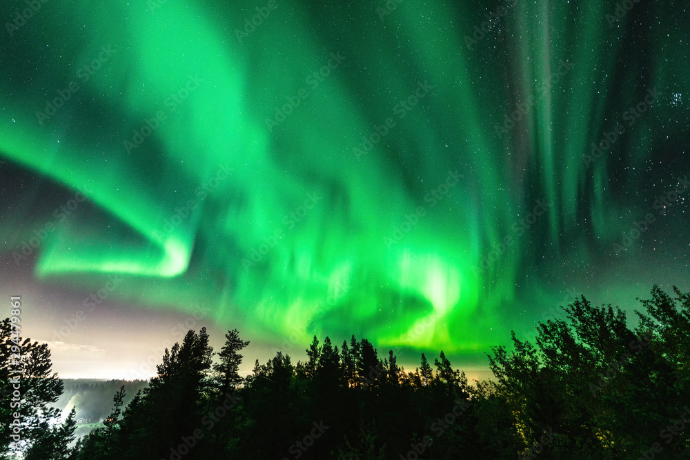 View of brilliant green Aurora shining over Swedish foggy forest landscape in mountains, light rays from a village and Northern Lights color sky in different soft colors, Northern Sweden, Scandinavia