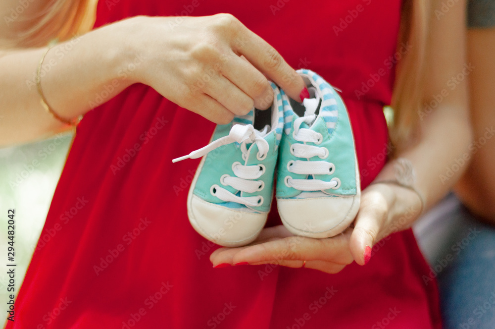 A happy married couple is holding blue booties in their hands. Husband hugs a beautiful adult pregnant wife. Second trimester. Waiting for a son.