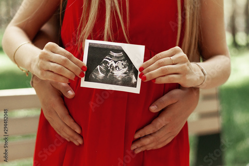 A happy married couple is holding in their hands a snapshot of an ultrasonic study of the baby. Husband hugs a beautiful adult pregnant wife. Second trimester.