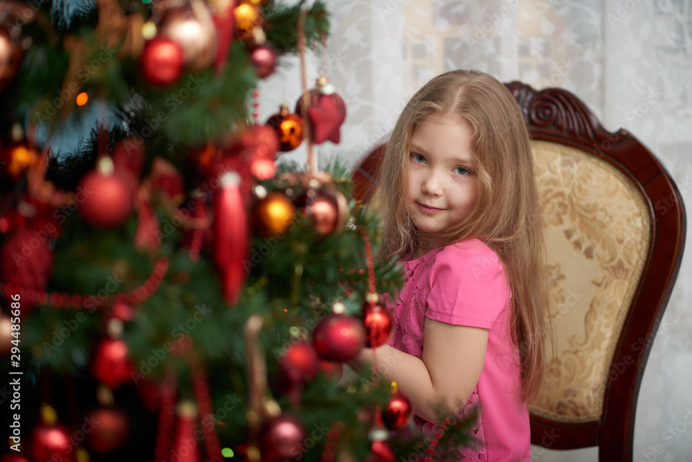 A little girl wearing light pink dress and  looking from behind a decorated Christmas tree.