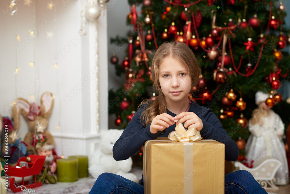 A young girl wearing a blue long sleeve shirt and jeans sitting on white carpet. Fireplace filled with toys and decorated Christmas tree background. A young girl wearing a blue long sleeve shirt and jeans sitting on white carpet. Fireplace filled with toys and decorated Christmas tree background.