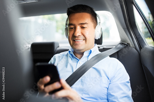 transport, business and technology concept - male passenger or businessman with wireless headphones using smartphone on back seat of taxi car