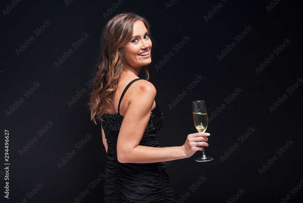 Young woman in elegant dress staying with glass of champagne against black background.