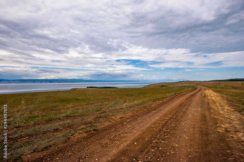 A dirt brown road stretches into the distance along the shore of a lake in cloudy weather. Behind the lake are mountains, the sky is overcast.