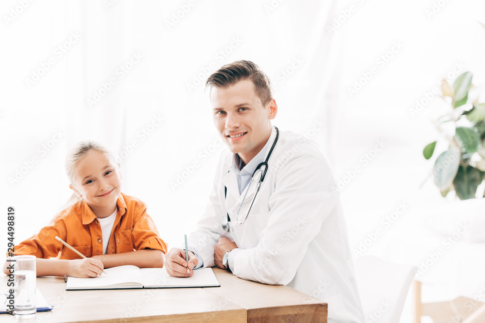 smiling child and pediatrist writing in notebook at table in clinic smiling child and pediatrist writing in notebook at table in clinic