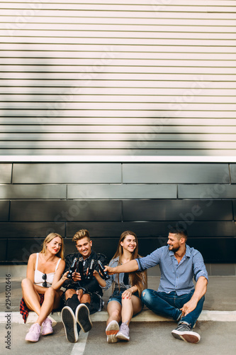 A group of stylish happy friends, girls and guys sit together in the city on the asphalt at the black wall, having fun and drinking drinks