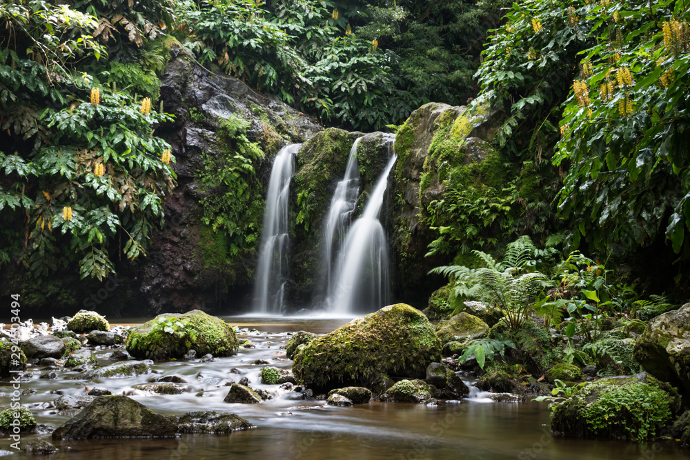 Natural waterfall surrounded by rocks and foliage at the Parque Natural da Ribeira dos Caldeirões, São Miguel island, Azores. Rocks in the meandering river are in the foreground. Long exposure image.