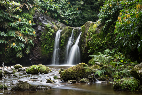 Natural waterfall surrounded by rocks and foliage at the Parque Natural da Ribeira dos Caldeirões, São Miguel island, Azores. Rocks in the meandering river are in the foreground. Long exposure image. photo