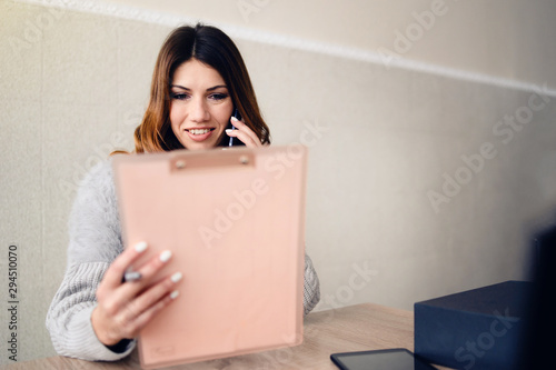 Young woman sitting by the table hold clip folder wearing formal talking to the phone while looking at clip pad paper documents at home or the office working