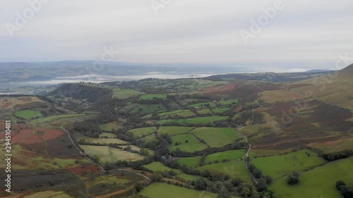An aerial backward footage of a Welch valley at dusk with green pasture, trees, rolling hills under a cloudy and misty sky 